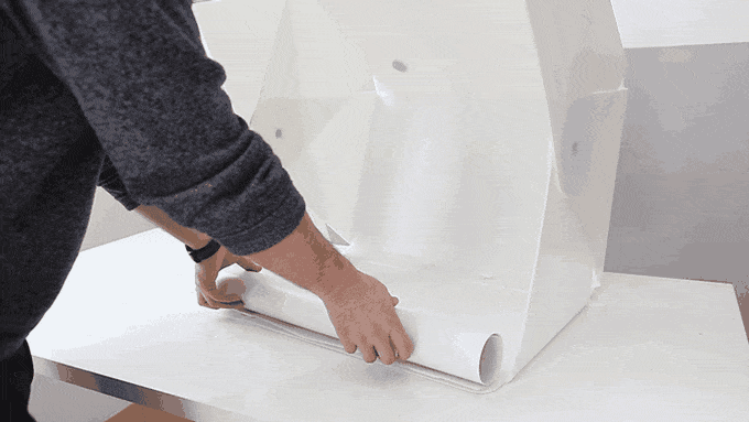 Eine Person baut einen weißen Leuchtkasten zusammen, indem sie Klettbänder auf einer flachen Oberfläche ausbreitet und anbringt. Der Leuchtkasten nimmt allmählich Gestalt an, während die Person das Material glättet und sicherstellt, dass es sicher befestigt ist.