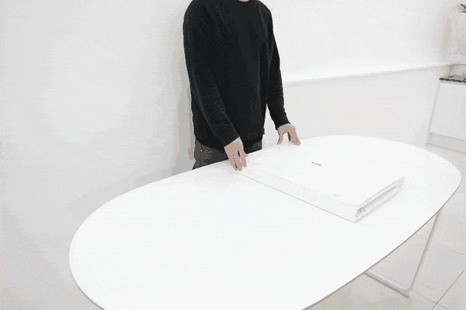 Eine Person in einem schwarzen Pullover demonstriert auf einem weißen, ovalen Tisch in einem gut beleuchteten Raum mit weißem Hintergrund, wie man eine Fotobox aufbaut.