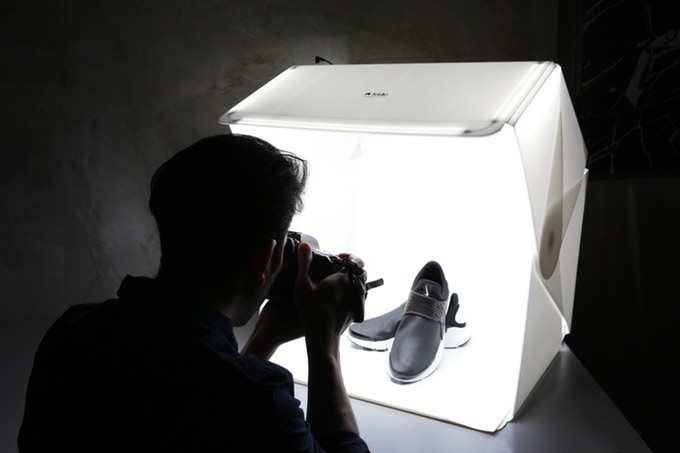 Eine Person fotografiert ein Paar schwarze Schuhe in einem Leuchtkasten. Der Leuchtkasten strahlt helles weißes Licht aus und beleuchtet die Schuhe für ein Foto in professioneller Qualität. Der Hintergrund der Szene ist dunkel und unscharf.