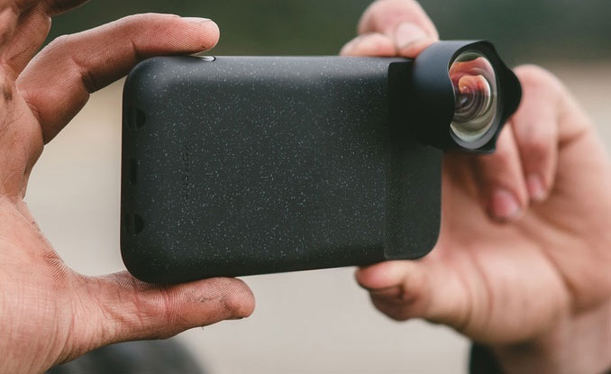 Eine Person hält ein Smartphone mit einer schwarz gesprenkelten Hülle, an deren Kamera ein Weitwinkelobjektiv angebracht ist. Der Hintergrund ist unscharf, wodurch das Telefon und das Objektivzubehör hervorgehoben werden.