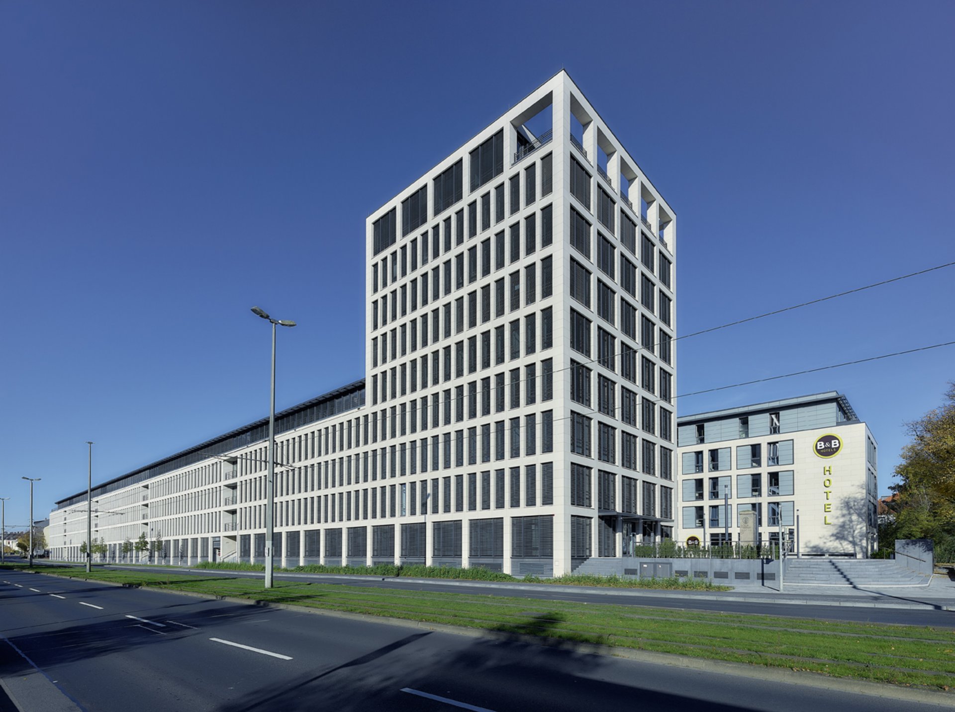 Ein modernes, mehrstöckiges Gebäude mit einer gitterartigen Fassade und großen Fenstern, gelegen neben einer Straße und einem grünen Rasen.