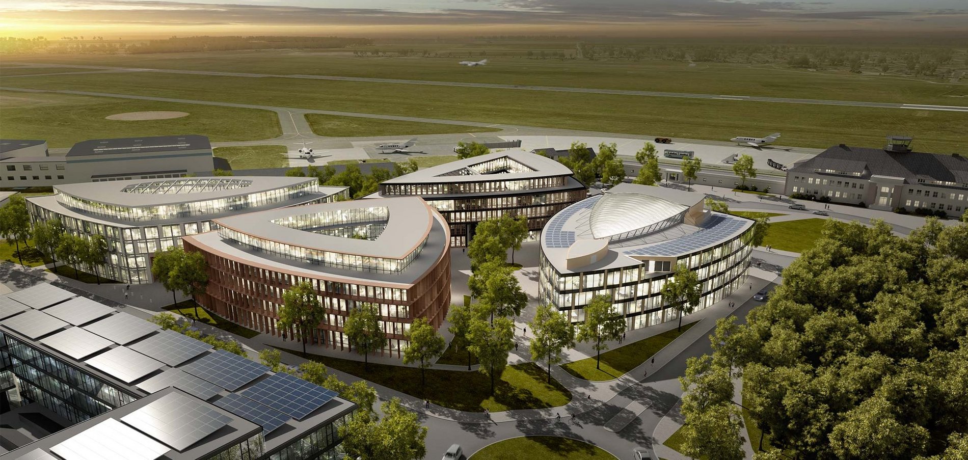 Luftaufnahme eines modernen Architekturkomplexes mit mehreren einzigartig geformten Gebäuden, umgeben von Grün. Der Komplex liegt neben einer Landebahn, auf der kleine Flugzeuge zu sehen sind, und am Horizont ist bei Sonnenaufgang oder Sonnenuntergang ein bewölkter Himmel zu sehen.